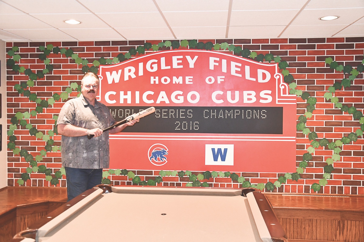 Bob Ibach’s sports memorabilia collection is extensive. Here he’s posed in front of his handpainted wall of the Cubs Ivy by Cathy Weadley. (Photo by Christine Such/Sun Day)